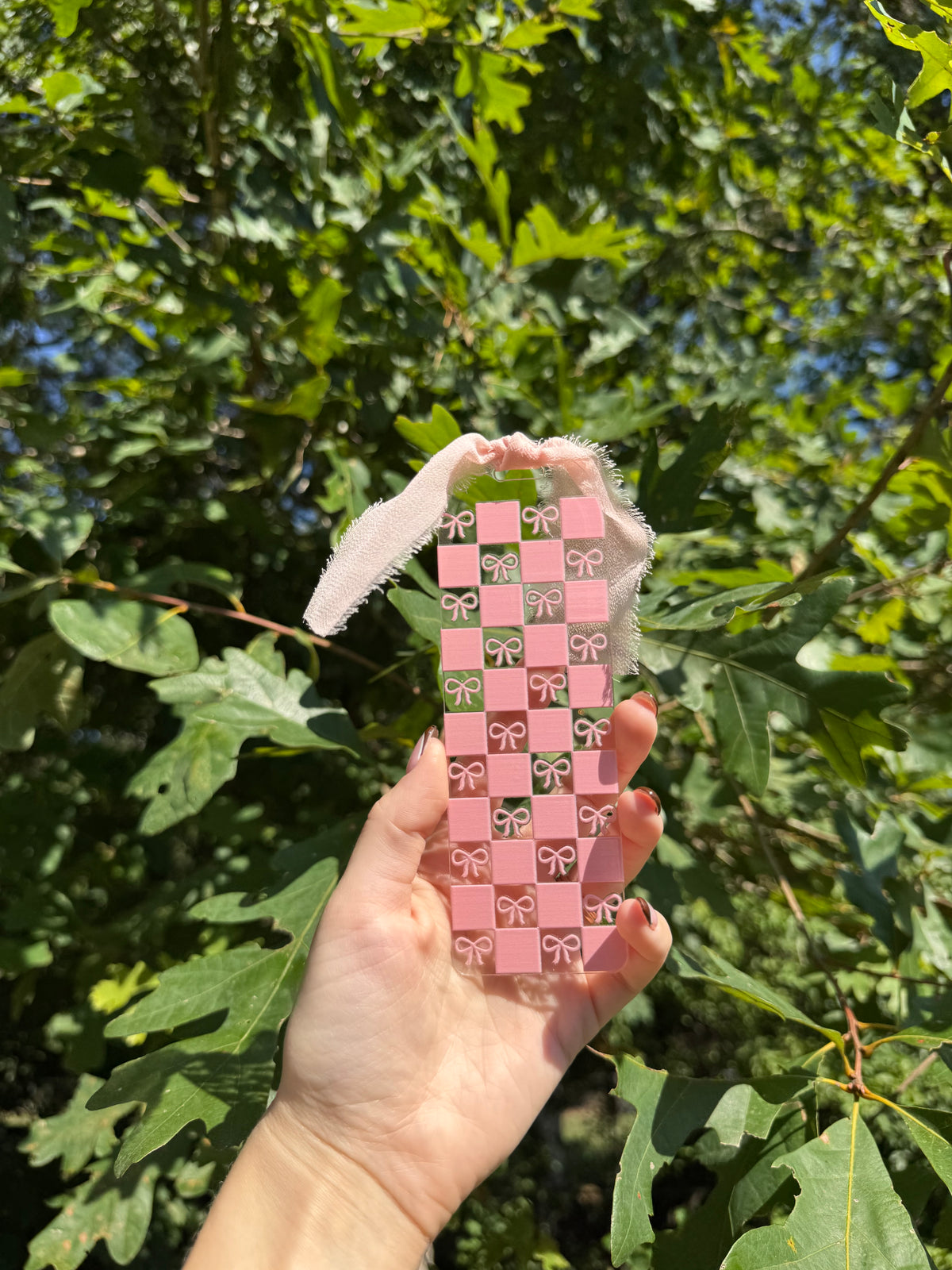 Pink checkerboard and bows acrylic bookmark with blush ribbon held outdoors