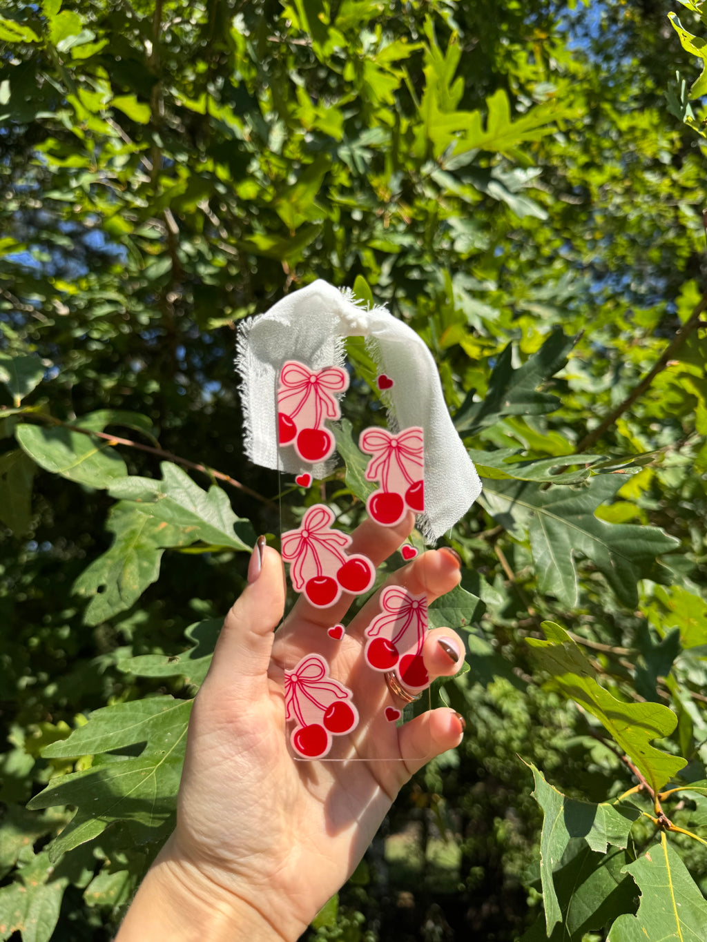 Clear acrylic bookmark with red cherries, pink bows, and white ribbon held outdoors