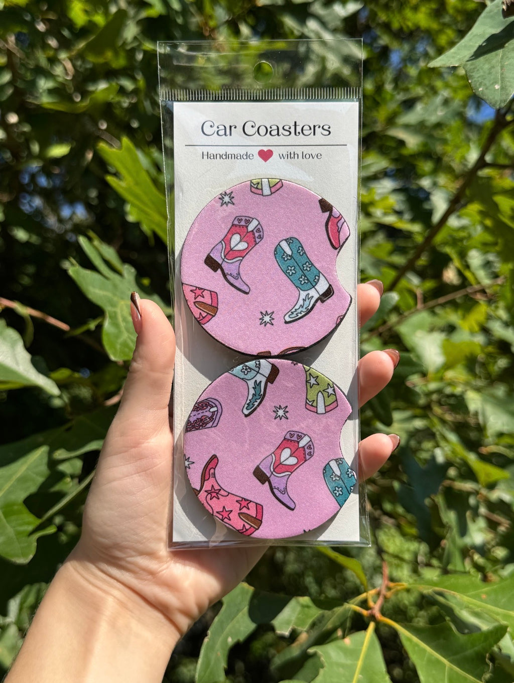 Set of two pink cowboy-boot car coasters in packaging against leafy trees